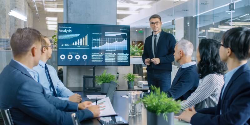 A Stock Trader Shows the Correlation among Cryptocurrency and Trade Market to his team pointing at the Wall TV.